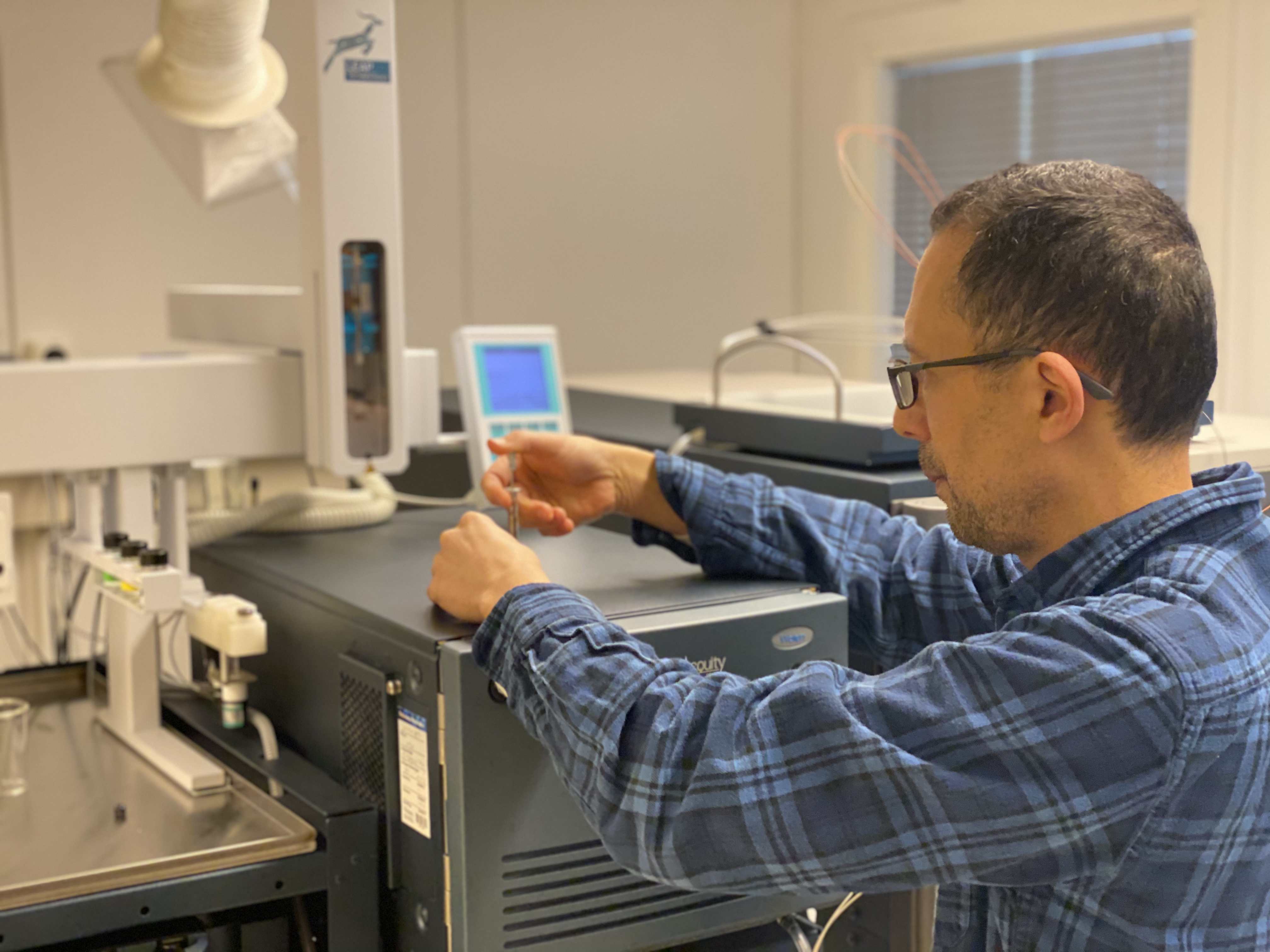 Dario Segura Peña wearing a blue plaid shirt and glasses and holding a syringe on top of the mass spectrometry instrument