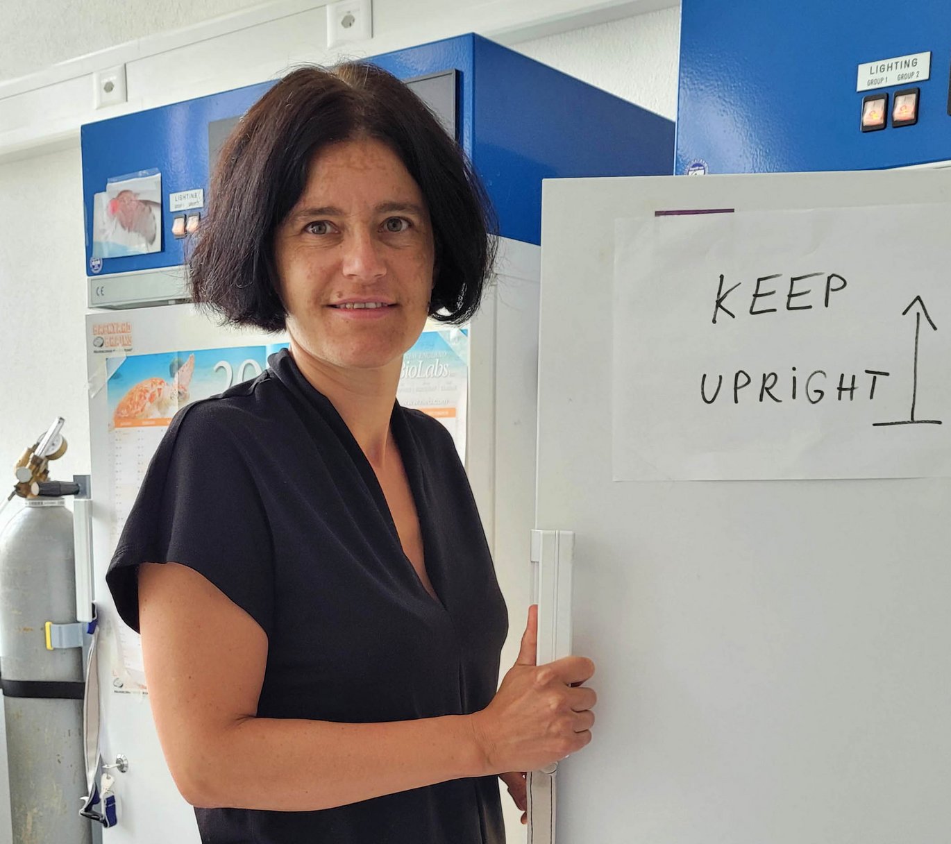 Prof. von Philipsborn wearing a dark shirt standing at a lab freezer with a sign “KEEP UPRIGHT” directing movers to keep the freezer standing during the move.