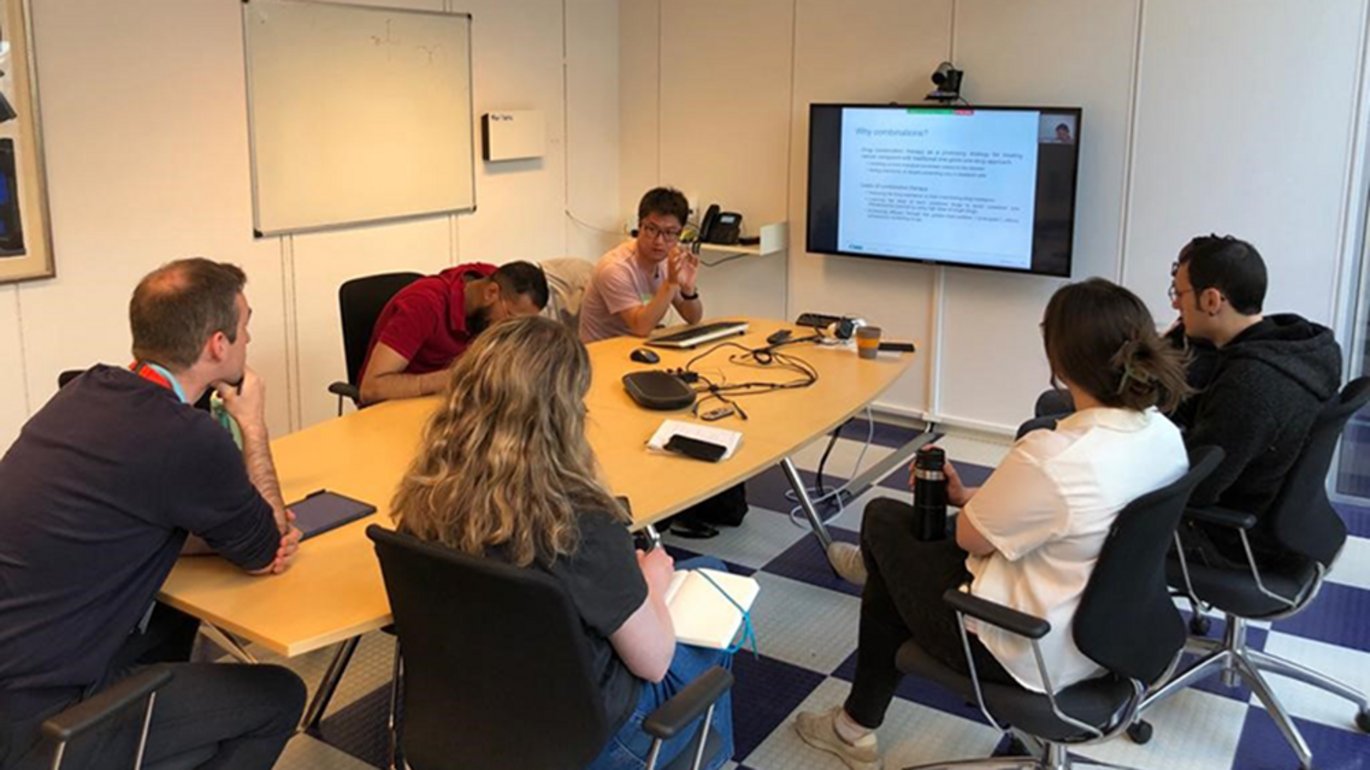 six people sitting in a room around a table looking slides projected onto a screen