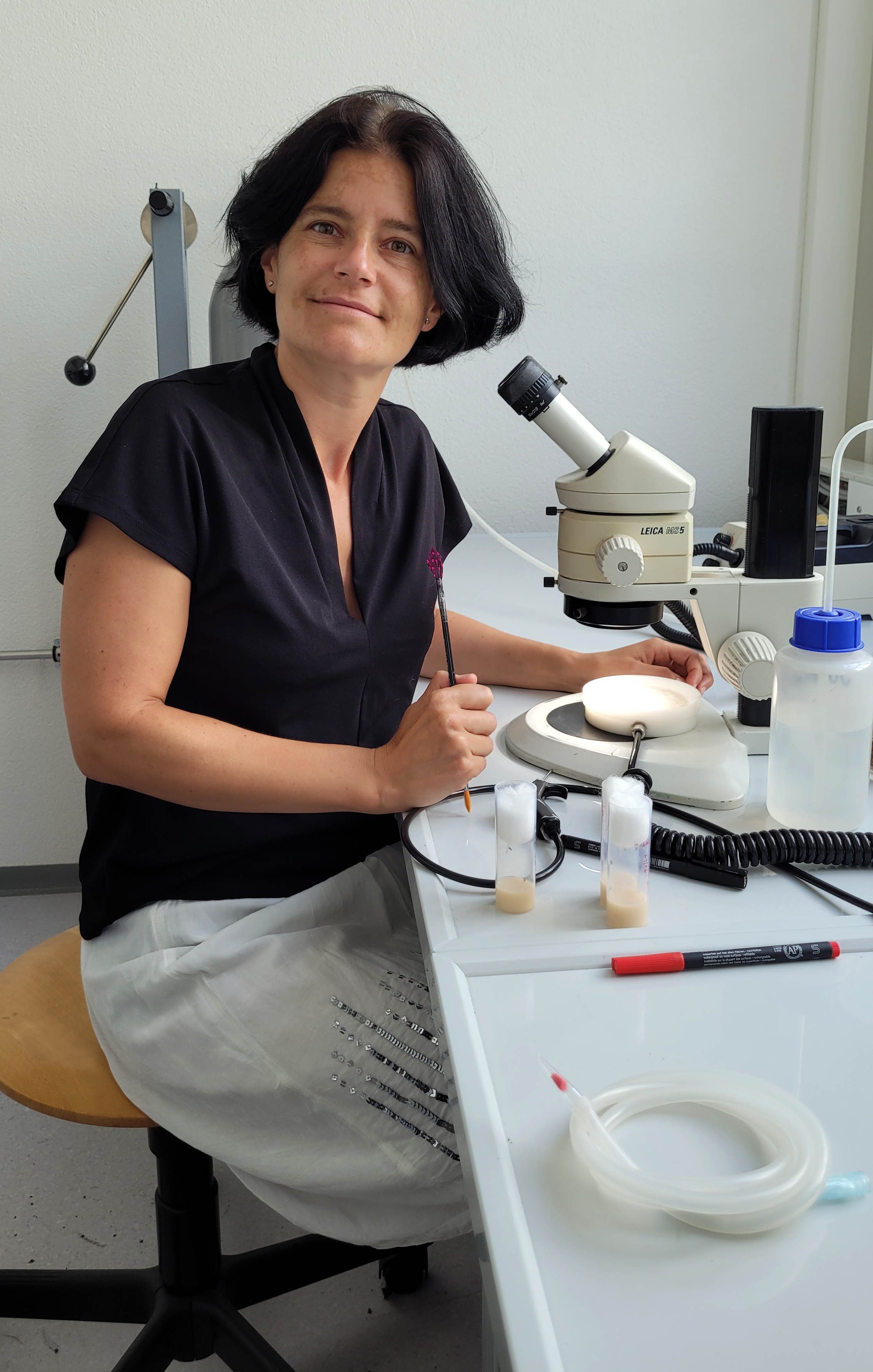 Prof. von Philipsborn wearing a dark shirt and light skirt sitting at a low table with lab supplies on it.