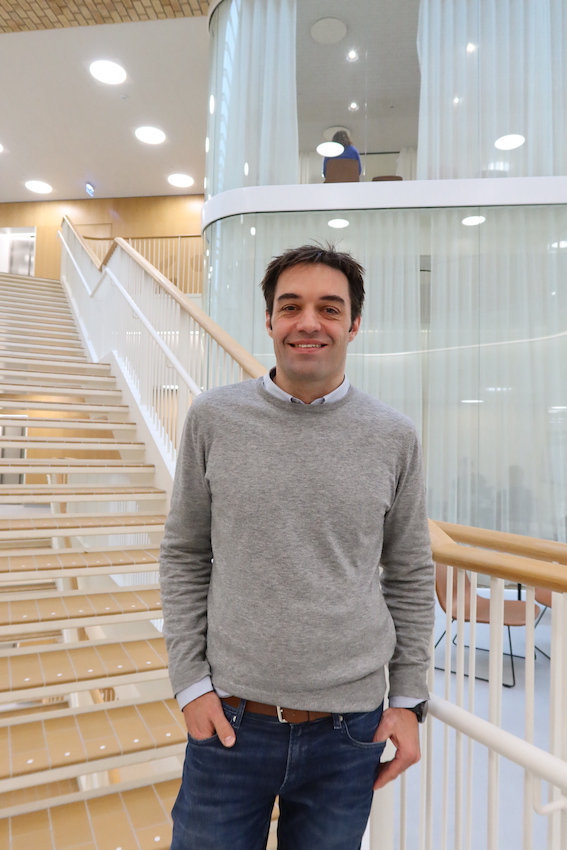 Mark Denham standing at the bottom of a staircase wearing a gray sweater and blue jeans