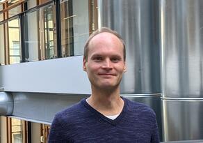 Dr. Paavolainen in a blue sweater with a background of the interior of the FIMM research building.